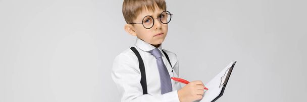 A charming boy in a white shirt, suspenders, a tie and light jeans stands on a gray background. The boy holds a pen and sheets for notes - Foto, immagini