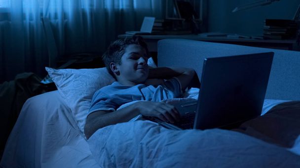 Boy chatting with friends on laptop in bed instead of sleeping, playing games - Foto, immagini