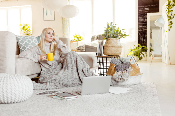 Beautiful young woman wearing sweater sitting on carpet in living room covered with blanket and resting with cup of coffee. Winter time. - 写真・画像