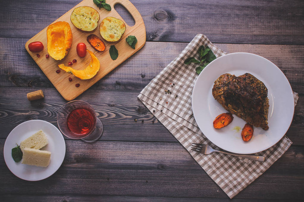A piece of meat, sprinkled with spices, baked with baked carrot slices on a white plate, fork, linen checkered napkin, next to the baked slices of pumpkin, potatoes, spices and cherry tomatoes. - Фото, изображение