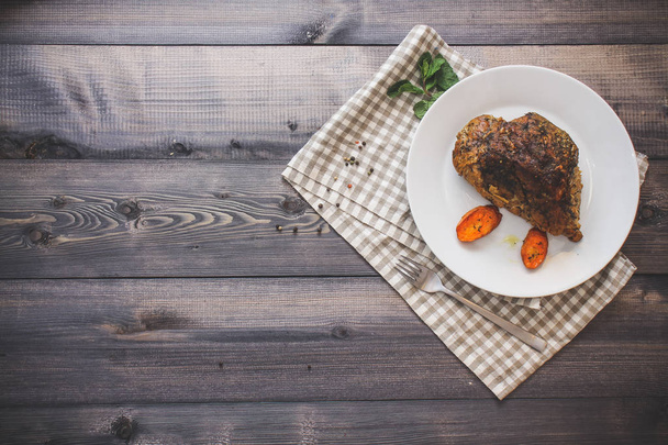 A piece of baked meat and slices of baked carrots on a white plate on a linen napkin in a cage on a dark gray tabletop - 写真・画像