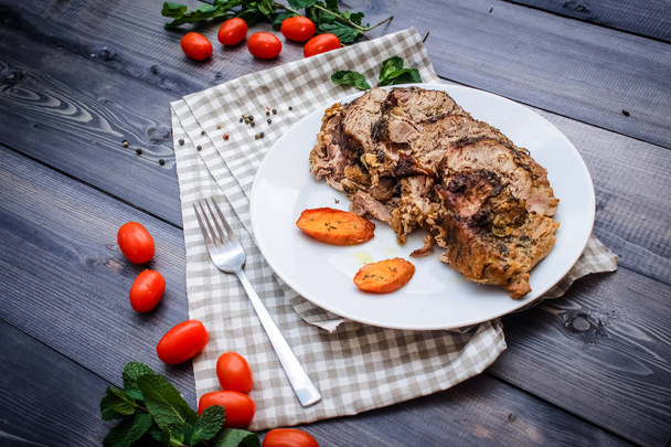 Chopped piece of meat baked in spices, baked carrot slices and greens on a white plate on a checkered linen napkin, surrounded by cherry tomatoes and greens around a napkin on a dark gray wooden table top - Fotografie, Obrázek