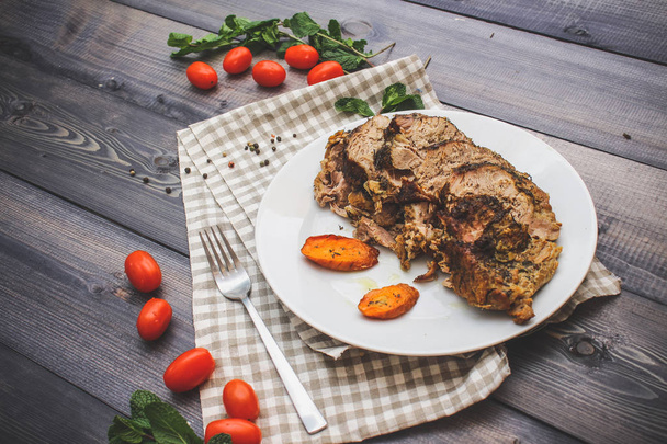 Chopped piece of meat baked in spices, baked carrot slices and greens on a white plate on a checkered linen napkin, surrounded by cherry tomatoes and greens around a napkin on a dark gray wooden table top - Foto, Imagem
