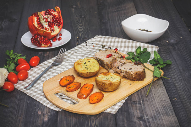 On the cutting board are two slices of baked meat, two slices of potatoes and baked carrot slices, a checkered napkin of flax, in a bowl are multi-colored peas of pepper and a cut pomegranate on a plate against the graphite tabletop - Foto, Bild