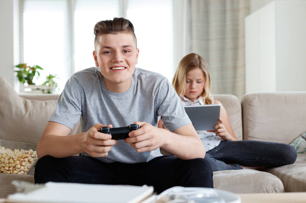 Brother and sister spending time at home, sitting on sofa in living room. Boy playing video game and girl using digital tablet.  - Foto, afbeelding