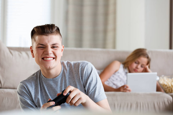 Brother and sister spending time at home, sitting on sofa in living room. Boy playing video game and girl using digital tablet.  - Foto, Imagem