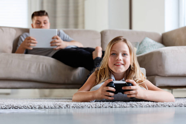 Brother and sister spending time at home. Girl laying on floor and playing video game while boy sitting on sofa and using digital tablet.  - Fotografie, Obrázek