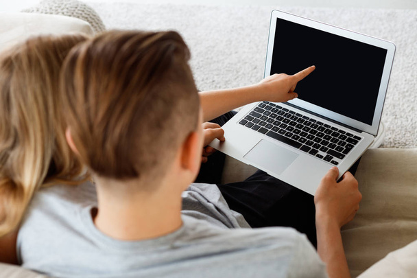 Back view of boy and girl sitting on sofa at home and using laptop. Girl pointing at computer screen. Unrecognizable people.  - 写真・画像