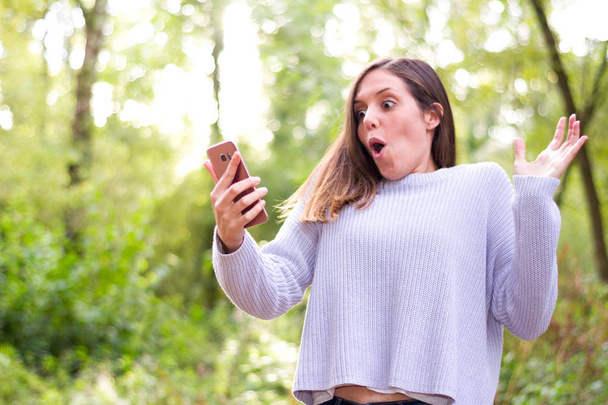 Europese jonge brunette vrouw zeer verbaasd op zoek naar de nieuwe mobil-vacatures in natuur of forest - Foto, afbeelding