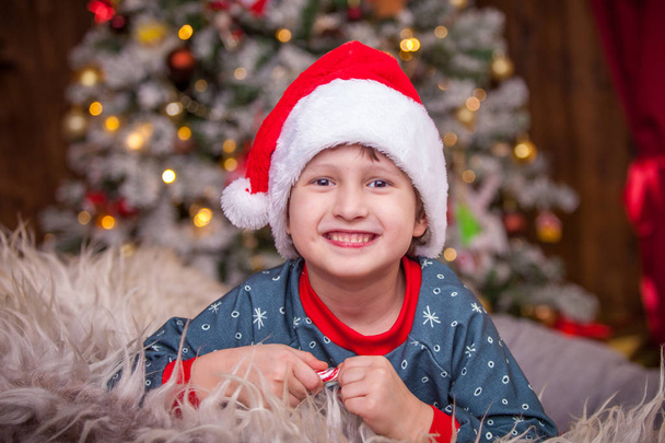 広いサンタ帽子の男の子の赤ちゃんが笑っています。部屋はすべてライトのクリスマス ライト飾られています。エレガントな雪に覆われたクリスマス ツリー フォーカス。クリスマスは、子供を喜ばせる家族の休日 - 写真・画像