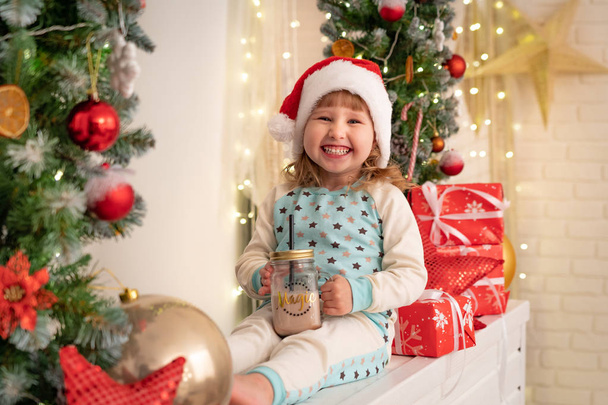 little girl in pajamas and Santa hat drinking cocoa milk! Christmas atmosphere surrounds. boxes of gifts tied with satin ribbons. the Christmas tree garland is decorated with dried oranges and balls - 写真・画像