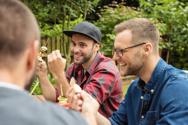 Outdoor shot of three friends enjoying barbecue party in garden, sitting at the table and eating vegetable skewers. - Foto, Imagen