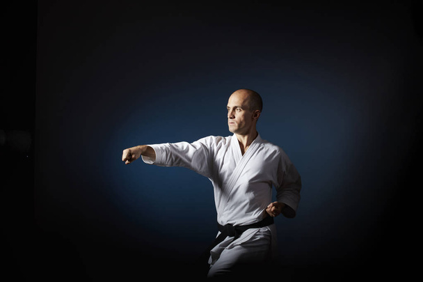 Op een blauwe achtergrond met een kleurverloop doet een atleet met een zwarte gordel formele karate oefeningen. - Foto, afbeelding