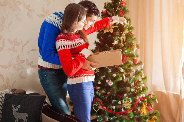 Couple in love decorating Christmas tree with toys at home, wearing winter sweaters. Preparing to New year - Photo, Image