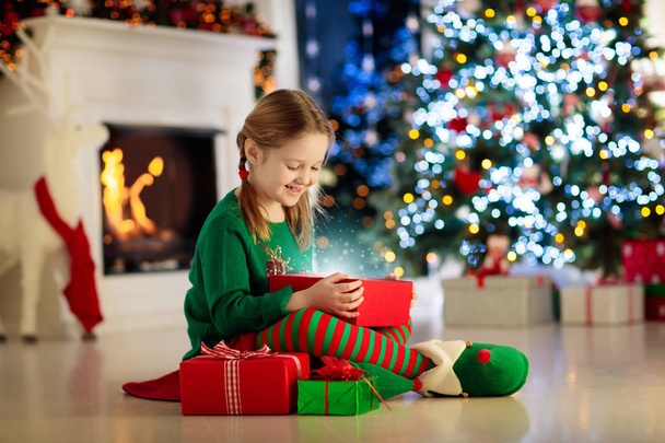 Child opening present at Christmas tree at home. Kid in elf costume with Xmas gifts and toys. Little girl with gift box and candy at fireplace. Family celebrating winter holidays. Home decoration. - Фото, зображення
