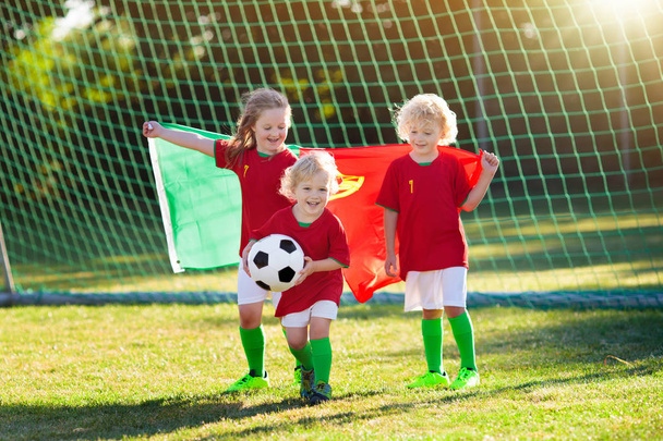 Τα παιδιά παίζουν ποδόσφαιρο σε υπαίθριο γήπεδο. φίλαθλοι της ομάδας της Πορτογαλίας με εθνική σημαία. Τα παιδιά σκοράρουν ένα γκολ στο ποδόσφαιρο. Παιδί με Πορτογαλική φανέλα και μπάλλα που κλωτσάνε. Θαυμαστής γιορτάζει νίκη στο γήπεδο. - Φωτογραφία, εικόνα