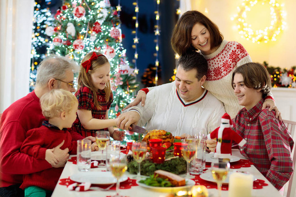 Gezin met kinderen die kerstdiner bij de open haard en versierde kerstboom eten. Ouders, grootouders en kinderen op feestmaal. Wintervakantie feest en eten. Kinderen openen cadeautjes en cadeaus. - Foto, afbeelding