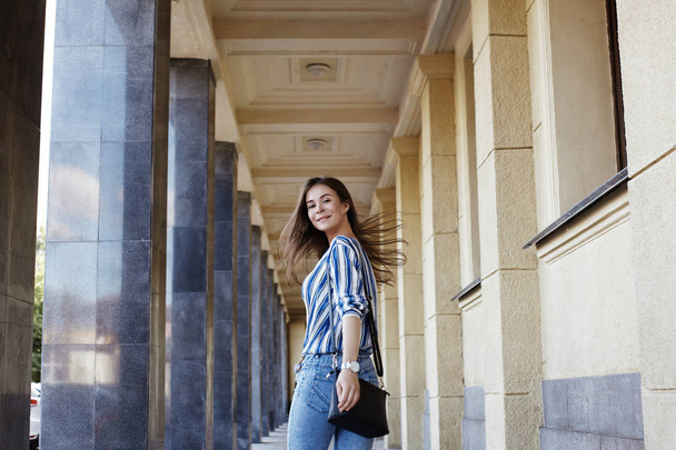 Street Style Outdoors Portrait of Beautiful Girl. Young Woman Smiling. She wearing Print Shirt, Blue Jeans and Black Bag. Happy Lifestyle shoot - Zdjęcie, obraz