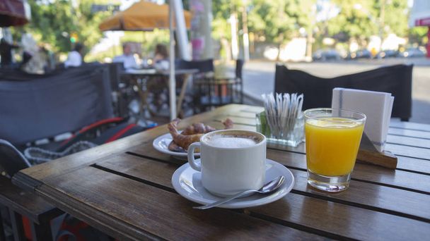 White cup with coffee with milk and glass of orange juice on a wooden table outside of a restaurant on a sunny morning. Breakfast photo - Photo, Image