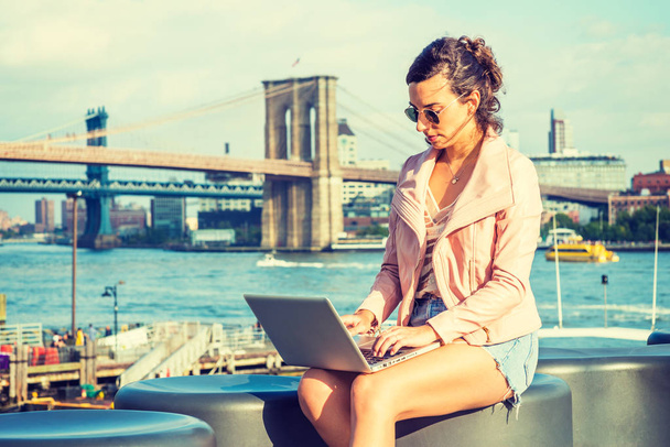Młoda Amerykanka podróże, praca w Nowym Jorku, noszenie różowa skórzana kurtka, niebieski Denim szorty, Okulary przeciwsłoneczne, siedzi nad rzeką, pracę na komputerze przenośnym. Brooklyn, Manhattan bridges w tle - Zdjęcie, obraz