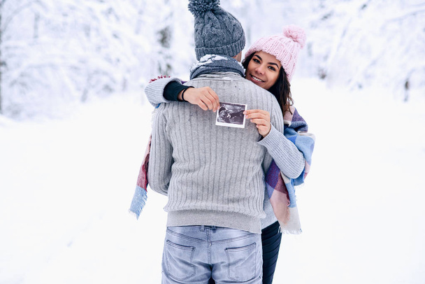 Beautiful pregnant woman in warm pink hat hugs gently her husband and holds ultrasonic pregnant picture in her hand. Family love and care concept. - Foto, afbeelding