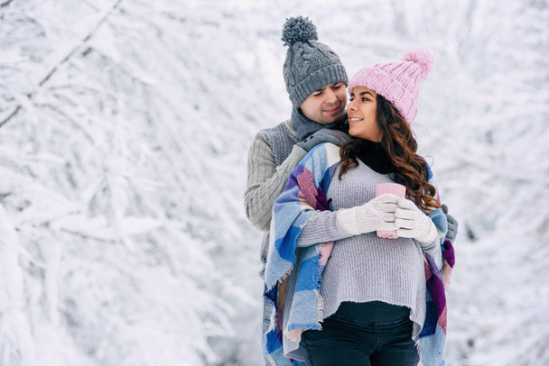Beautiful pregnant woman with her husband dressed grey knitted sweaters, hat, scarf and jeans, holding pink cups with hot tea and walking in a snowy winter park on background. - Фото, зображення