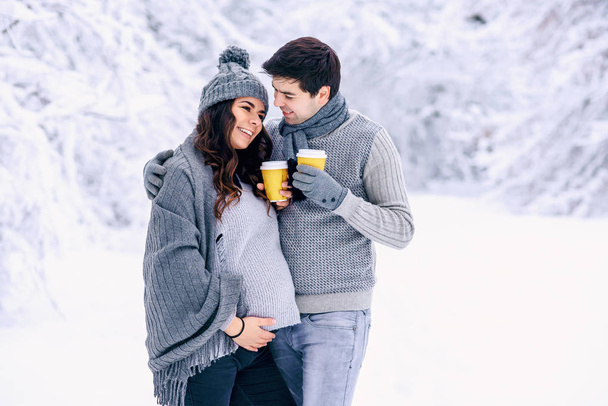 Beautiful pregnant woman with her husband dressed grey knitted sweaters, hat, scarf and jeans, holding yellow cups with hot tea and walking in a snowy winter park on background. - Foto, Imagem