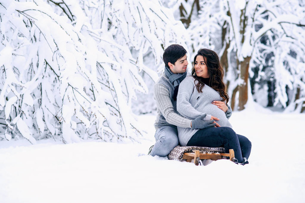 Beautiful pregnant woman with her husband sitting on the sled in a snowy winter park. - Fotografie, Obrázek