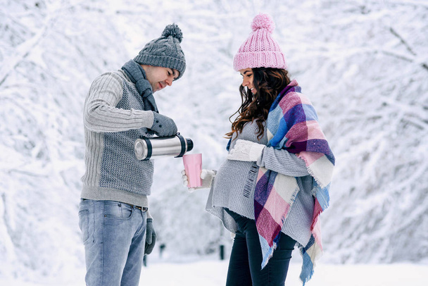 Beautiful pregnant woman with her husband walks in the snowy park. A man pours warm tea for his lovely wife. - Foto, Imagen
