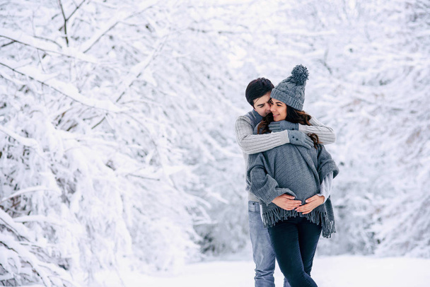 Husband in a grey sweater and jeans tenderly hugging his beautiful pregnant wife in grey knitted sweaters, hat, scarf and jeans in a snowy winter park. Pregnancy concept. - Photo, image