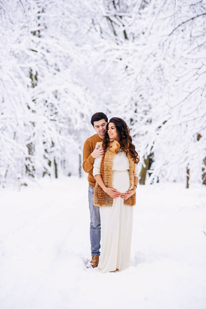 Husband in a sweater and jeans tenderly hugging his beautiful pregnant wife in a long light dress and coat in a snowy winter park. Family love and care concept. - Photo, Image