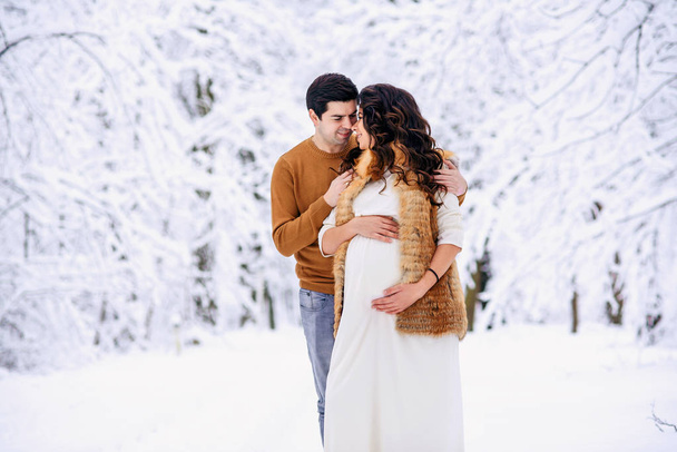 Husband in a sweater and jeans tenderly hugging his beautiful pregnant wife in a long light dress and coat in a snowy winter park. Family love and care concept. - Photo, Image