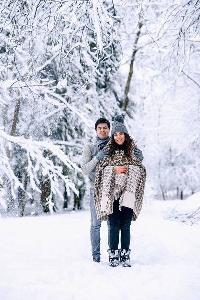 Young man hugs tenderly his beautiful pregnant wife covered in warm plaid. Couple in love walking together in snowy winter park. - Fotoğraf, Görsel