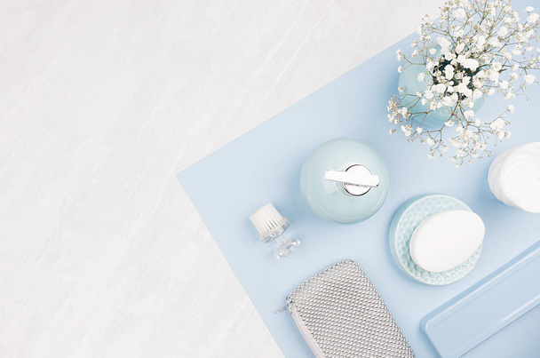 Fashion art cosmetics flat lay of different cosmetics products and accessories of silver, pastel blue and white color with small flowers on blue paper and white wooden background, copy space. - Фото, изображение