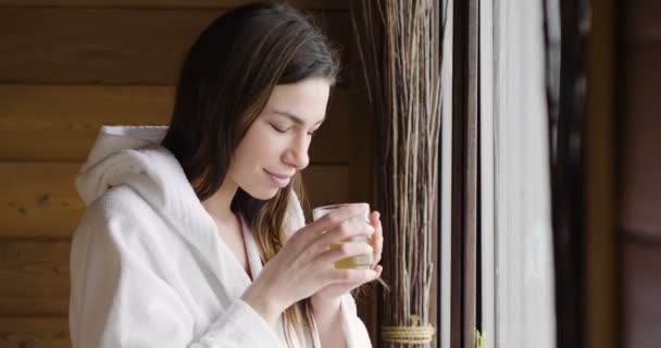 A beautiful woman looks out the window wearing a white bathrobe and drinks hot tea or herbal tea looking out. The woman relaxes and thinks as she looks out. - Кадры, видео