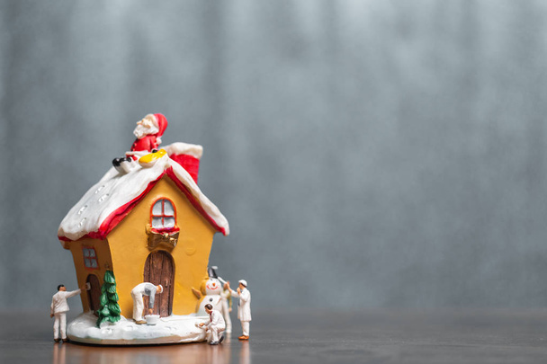 Μινιατούρα ανθρώπους ζωγραφική σπίτι και τον Άγιο Βασίλη να κάθεται στην οροφή, καλά Χριστούγεννα και καλές γιορτές έννοια. - Φωτογραφία, εικόνα