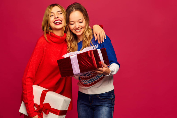 Karácsony, karácsonyi, koncepció. Két gyönyörű nők elegáns pulóverek mosolyogva. Lányok, pózol, piros háttér. Nagy ajándék dobozok modellek. Szórakozás, készen áll az ünneplésre. Fényes ünnep a legjobb barátai - Fotó, kép