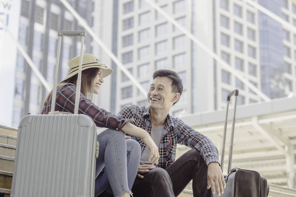 Nuori aasialainen pari matkailijoita Hymyilee onnellisesti istuessaan matkatavaroiden kanssa, käsite matkailijoiden rakastajan matkustamisesta etelä-aasialaisella taustapilvenpiirtäjällä.
 - Valokuva, kuva