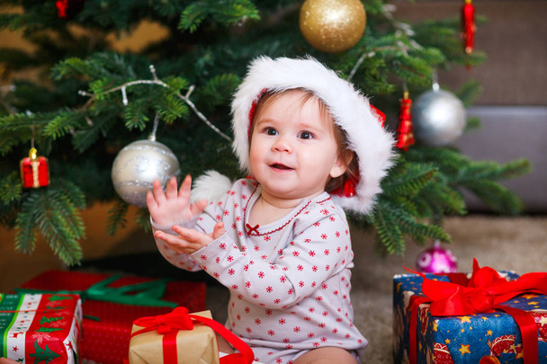 Little smiling baby in hat of Santa Claus on a background of Christmas garland, tree and gift boxes with ribbon. - Фото, изображение