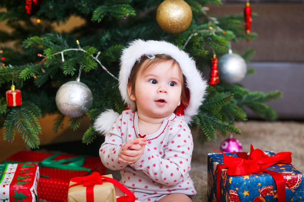 Little smiling baby in hat of Santa Claus on a background of Christmas garland, tree and gift boxes with ribbon. - 写真・画像