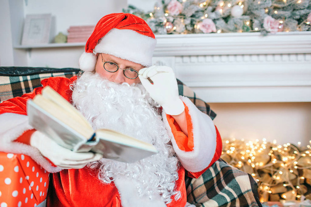 Serious and concentrated man in santa claus costume reads book. He holds hand on glasses. Guy pays attention to book. - Photo, Image