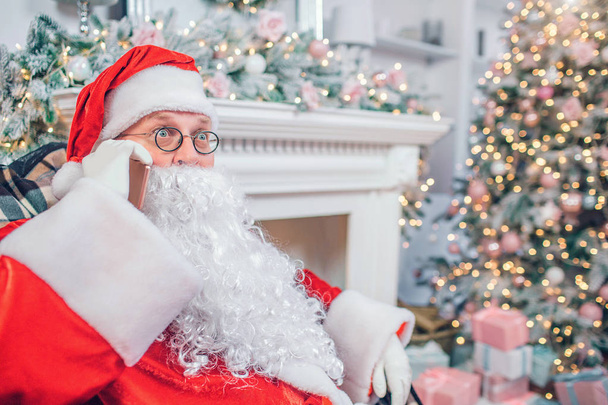 Wondered Santa Claus sits in festive room. He talks on phone. Guy is amazed. He is alone in room. - Foto, imagen