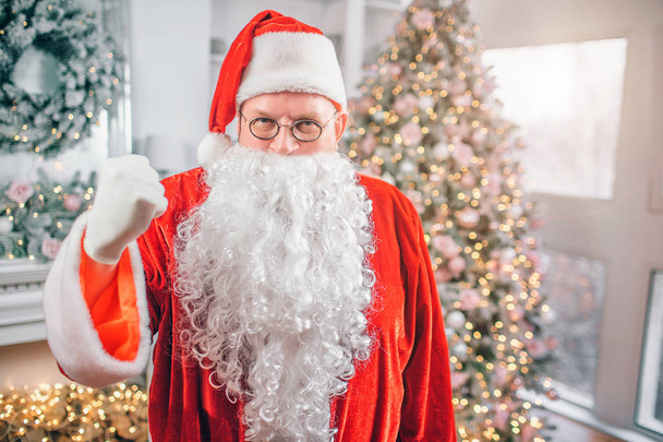 Picture of Santa Claus stands and looks on camera. He shows fist. Man threatens. He is in festive room. - 写真・画像