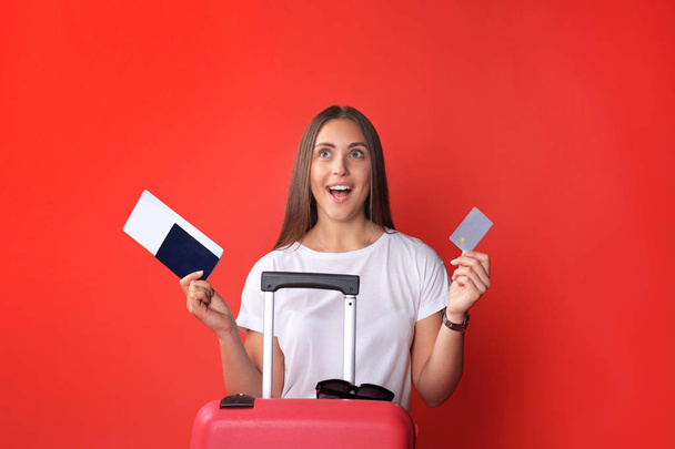 Tourist girl in summer showing plastic credit card, with sunglasses, red suitcase, passport isolated on red background. - Foto, Imagen