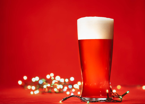 Pint glass of of pale lager beer or ale with big head of foam and christmas lights on red background - Photo, Image