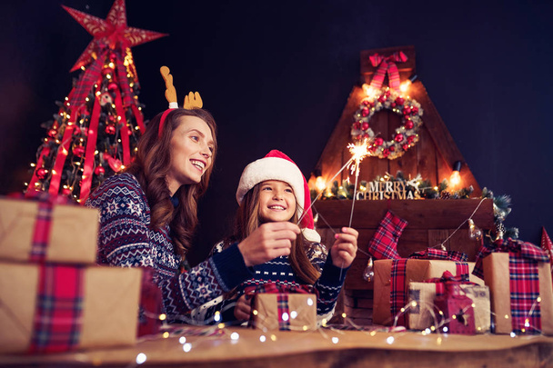 holidays, family and people concept. Happy mother and little girl in santa helper hat with sparklers in hands, gift boxes on foreground Having fun - Foto, immagini