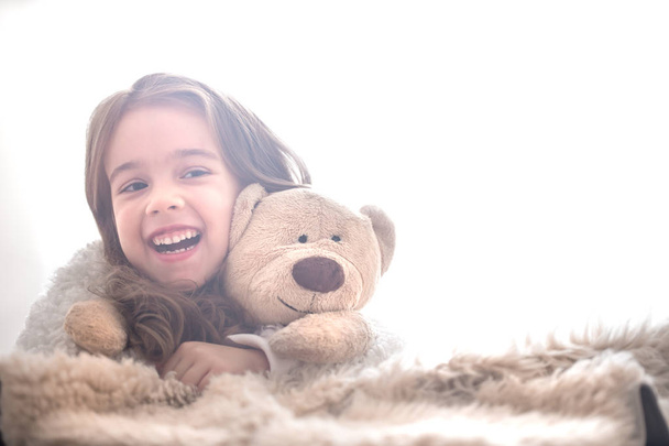 Маленька красива мила дівчинка обіймає іграшку плюшевого ведмедя на світлому тлі, місце для тексту
 - Фото, зображення