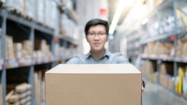 Young Asian man carrying cardboard box between row of shelves in warehouse. Shopping warehousing or working pick and packing concepts - Photo, Image