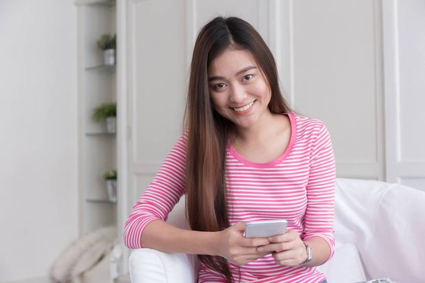 Ázsiai nő mosolyogva és smartphone segítségével elmosódott háttér: fehér hálószoba. - Fotó, kép