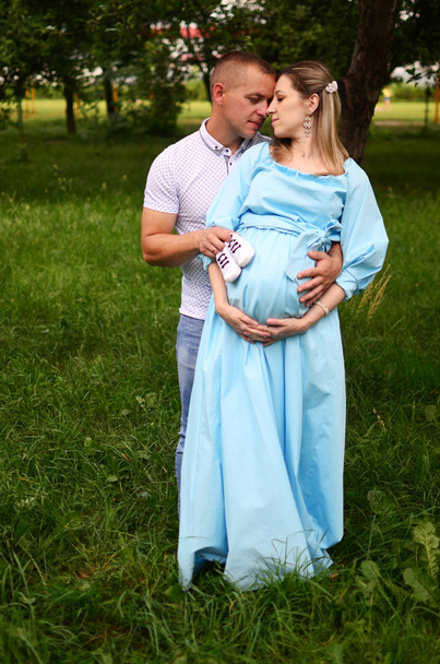 Happy family together in summer garden/park.Couple waiting for a baby. wife holding a pregnant belly. Husbent holding a tiny shoes. Outdoor photo - Photo, Image